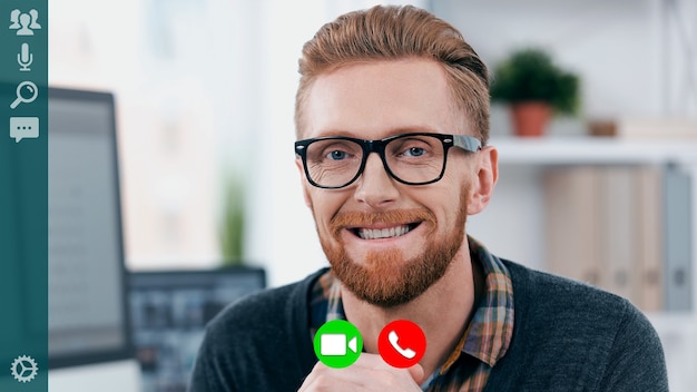 Handsome young man in smart casual wear looking at camera and smiling while having video call