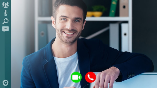 Handsome young man in smart casual wear looking at camera and smiling while having video call