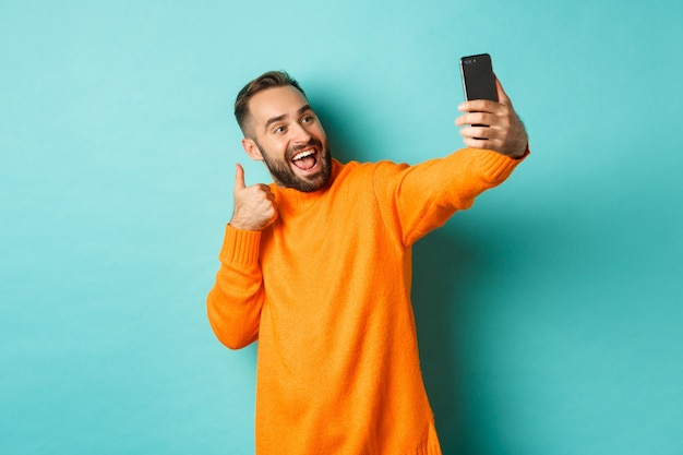 Handsome young man taking selfie on mobile phone, showing thumbs up at smartphone camera, recording