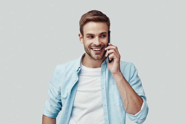 Handsome young man talking on the phone and smiling while standing against grey background