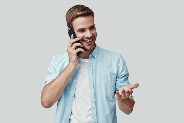 Handsome young man talking on the phone and smiling while standing against grey background