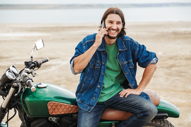 Handsome young man wearing casual outfit sitting on a motocycle at the beach, talking on mobile phone