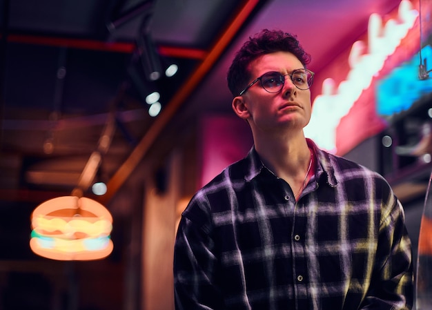 A handsome young man wearing a checkered shirt standing at night in the street. Illuminated signboards, neon, lights.