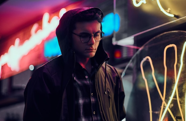 A handsome young man wearing sports hoodie standing in the street at night. Illuminated signboards, neon, lights.