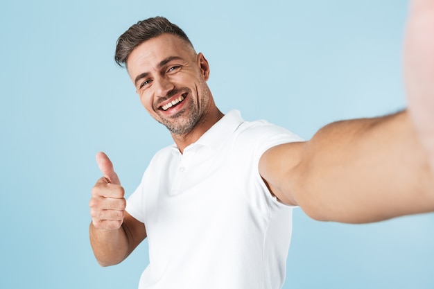 Handsome young man wearing white t-shirt standing over blue, taking a selfie, giving thumbs up