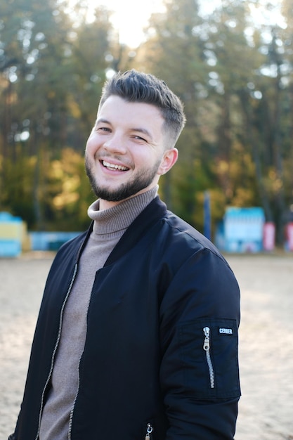 Handsome young man with a beard and in a black jacket posing on the beach Man smile and look at the camera