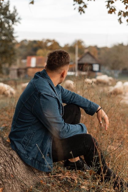 Handsome young man with hairstyle in a fashionable denim shirt and jeans sits and rest near a tree in the countryside and looks at sheep Guy travels in the countryside