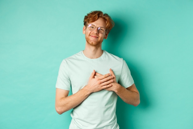 Handsome young man with red hair and glasses, holding hands on heart and smiling, saying thank you, feeling grateful and touched, standing over turquoise background