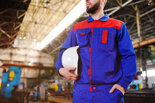 A handsome young man - a worker or an engineer with a white construction helmet in his hands against the backdrop of a plant or production