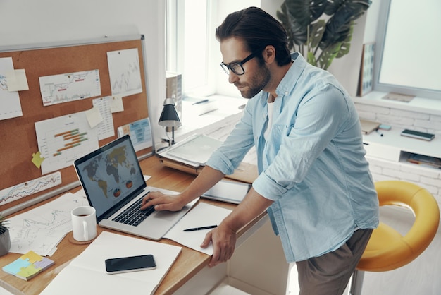 Handsome young man working on laptop while standing near his working place