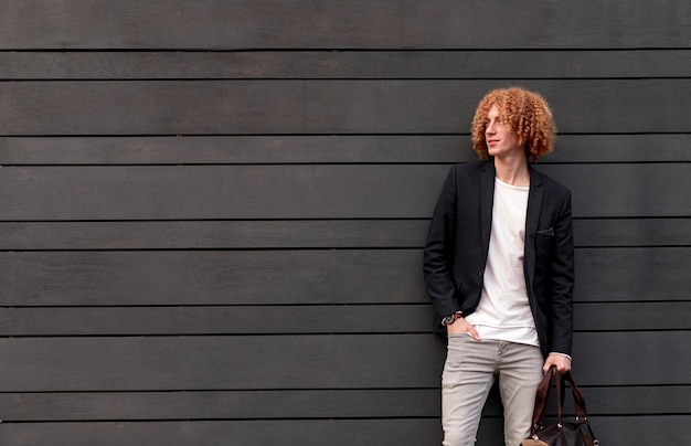 Photo handsome young self assured guy with red curly hair dressed in trendy outfit holding bag and looking away while standing against gray wall