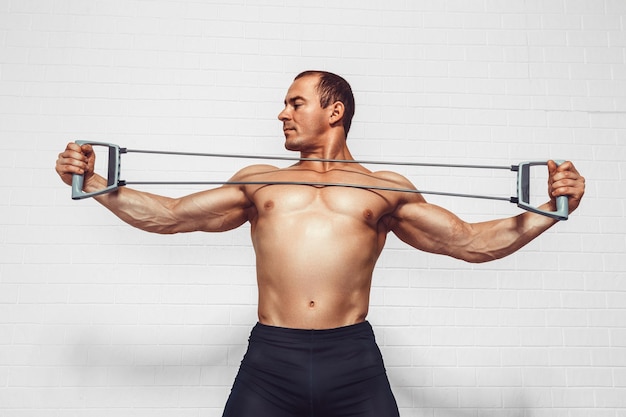 Handsome young sportsman exercising using expander while standing against white background