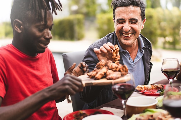 Happi multiethnic friends sitting at farmhouse table an African guy hands the grilled chicken wings to a mid aged man conviviality and friendship lifestyle concept