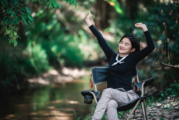 Happily young woman sitting on chair while relax time on camping in forest copy space