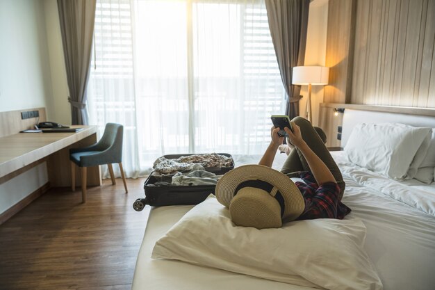 Happiness Asian traveler woman sleeping and using the smart mobile phoneon the bed in bedroom of hotel or hostel when traveling