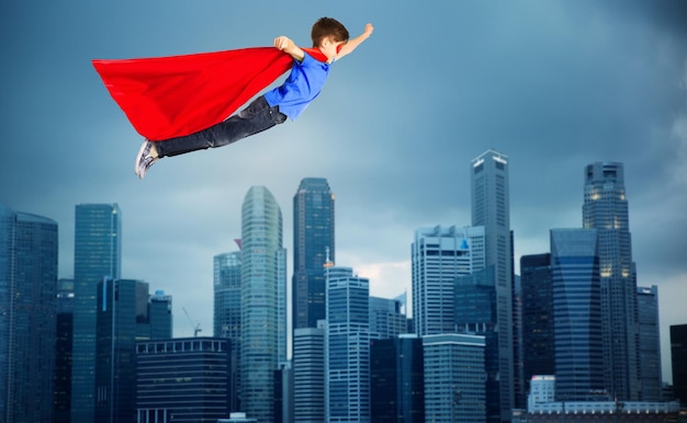 happiness, freedom, childhood, movement and people concept - boy in red superhero cape and mask flying in air over city background