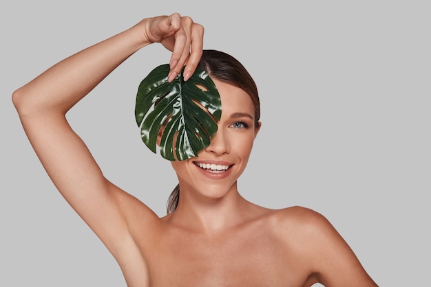 Happiness highlights her beauty. Attractive young woman looking at camera and covering face with leaf while standing against grey background