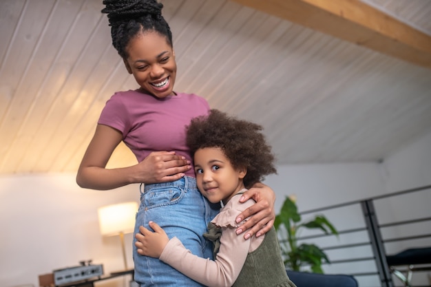 Happiness, life. Happy laughing dark skinned young woman and touching enthusiastic cute little daughter standing together at home