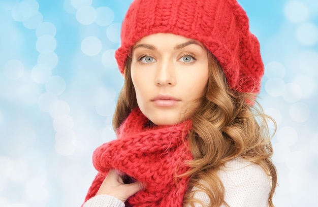 happiness, winter holidays, christmas and people concept - close up of young woman in red hat and scarf over blue lights background