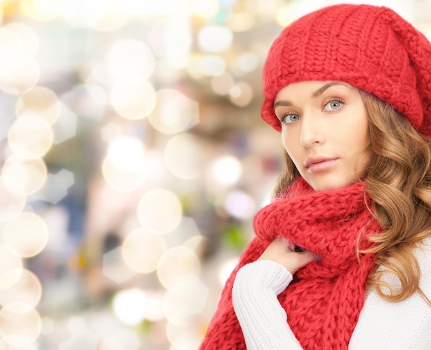 happiness, winter holidays, christmas and people concept - young woman in red hat and scarf over lights background