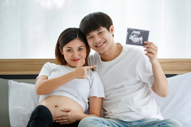 Happiness young Asian couple wearing a casual dress smiling and sitting on a bed together. A happy husband puts his arm around his pregnant wife and showing an ultrasound scan photo of a fetus.