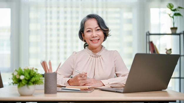 Happy 60s agedasian businesswoman sitting at her desk smiling and looking at the camera