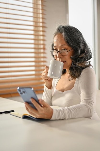 Happy 60s Asianaged woman sipping her morning coffee while using her phone at her desk