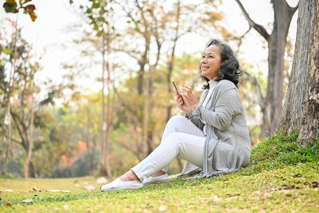 Happy 60yearold Asian woman using her phone while relaxing under a tree in a park