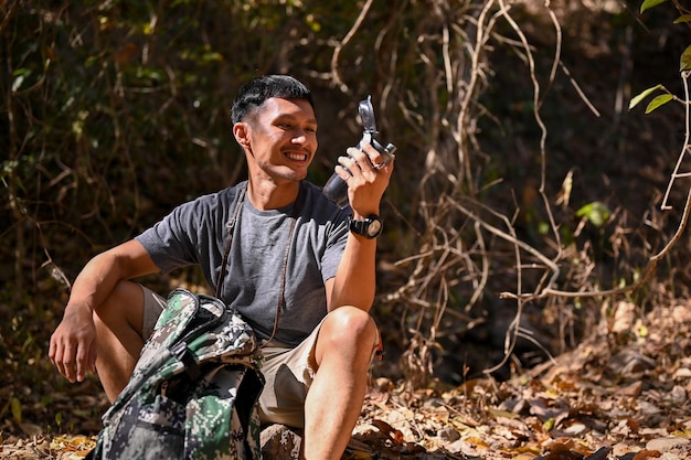 Happy and active Asian male traveler sits on a rock and drinks water from a bottle