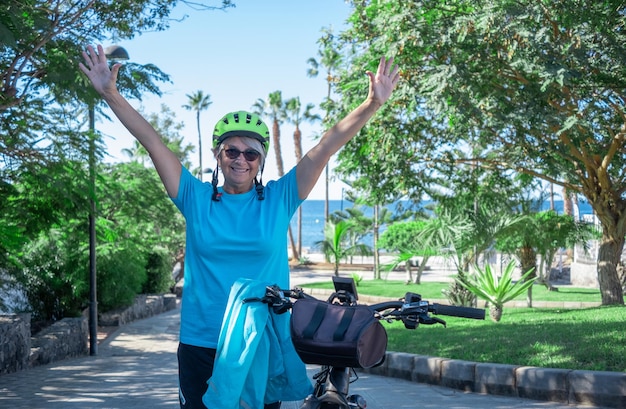 Happy active senior woman standing in the public park close to her electric bicycle enjoying freedom Sea on background Concept of healthy lifestyle and sustainable mobility