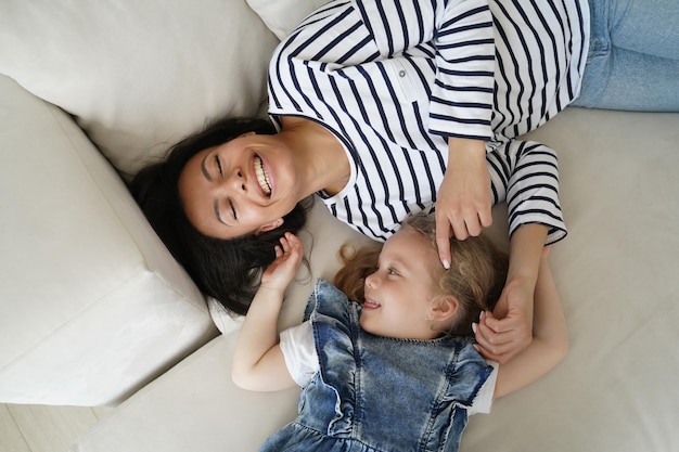 Happy adopted little kid girl tickling laughing mother lying having fun together on sofa top view