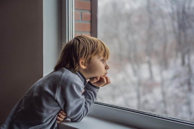 Happy adorable kid boy sitting near window and looking outside on snow on Christmas day or morning Smiling child fascinated with snowfall and big snowflakes