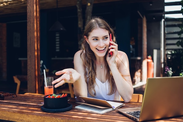 Happy adult female talking on smartphone sitting with laptop in lounge