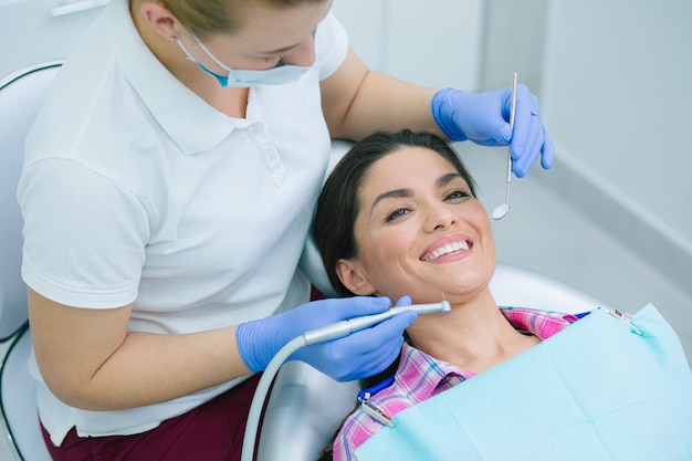 Happy adult woman smiling while a professional female dentist holding a mouth mirror and a high speed handpiece