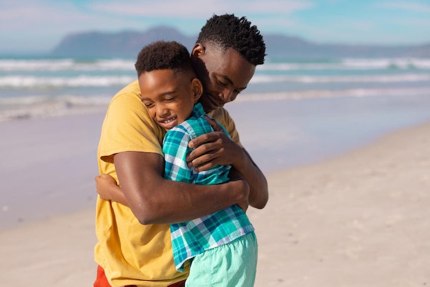 Happy african american boy with eyes closed embracing young father at beach against sea on sunny day