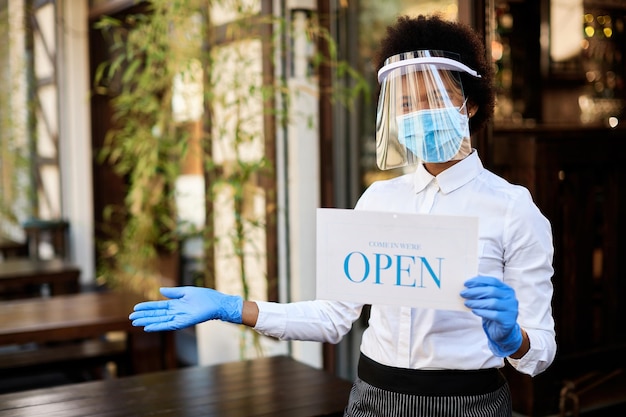 Happy African American cafe owner holding open sign while wearing visor and protective face mask