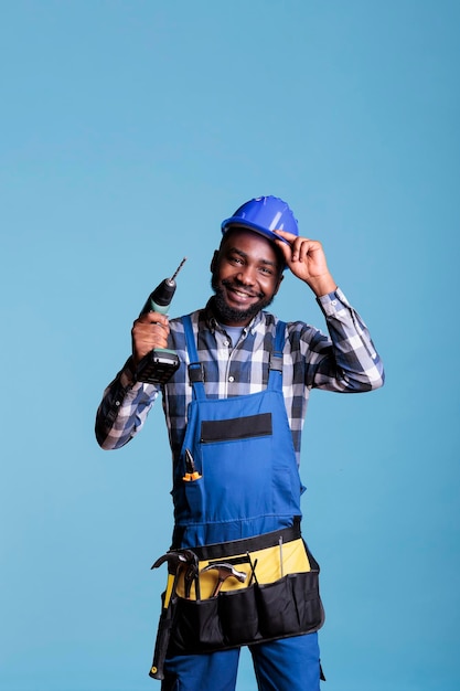 Happy african american construction worker adjusting hard hat holding electric cordless drill. Optimistic handyman wearing tool belt in studio shot isolated on blue background.