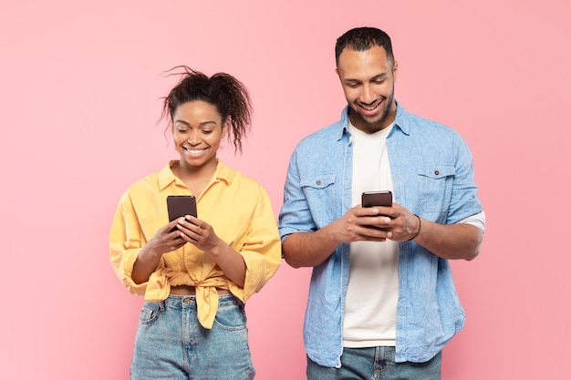 Happy african american couple holding their smartphones and surfing internet standing over pink studio background