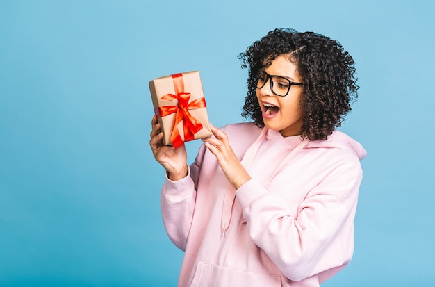 Happy african american curly lady in casual laughing while holding present gift box isolated over blue background.