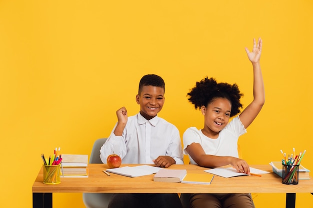 Happy african american female schoolgirl and mixed race schoolboy sitting together at desk and stretching arms up on yellow background Back to school concept