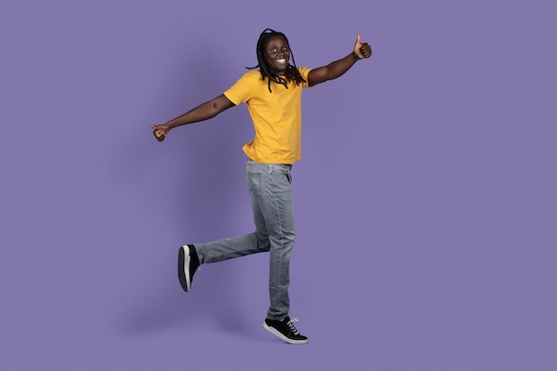 Happy african american guy jumping on purple showing thumb ups