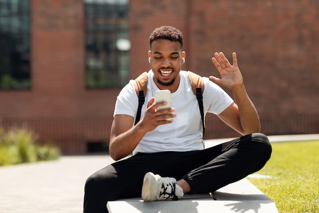 Happy african american guy making video call on cellphone waving hand to webcamera while walking in