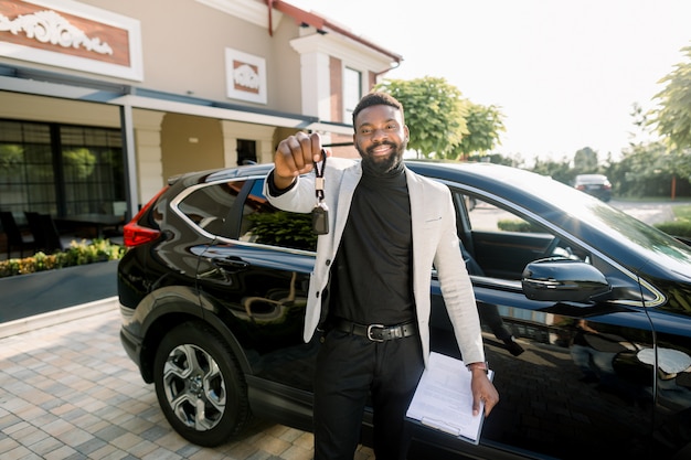 Happy African American man showing car key standing near new black car crossover outdoors. Young African American businessman holding a car key and car sales contract