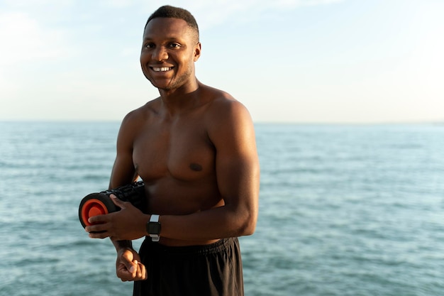 Happy african american man with naked torso standing at the seashore with yoga mat