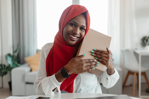 Happy african american muslim woman with red hijab got her delivery embracing box and smiling at camera