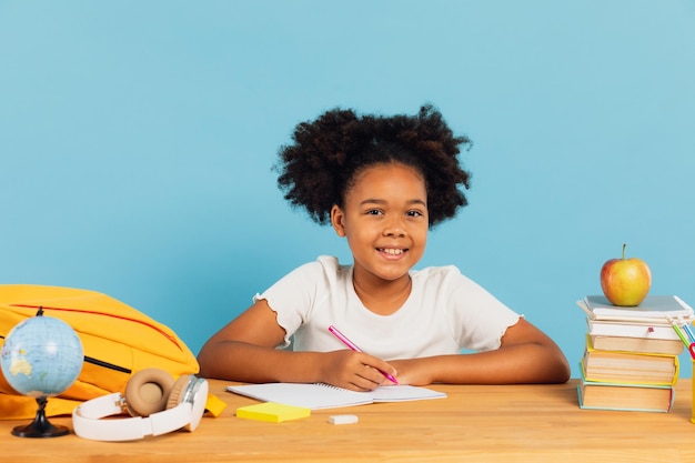 Happy African American schoolgirl doing homework at desk in class on blue background Back to school concept