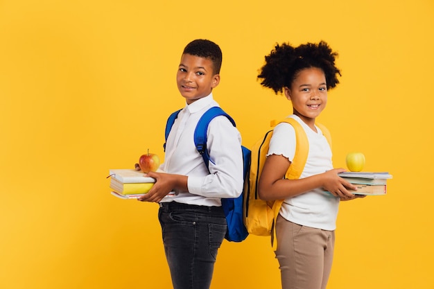 Happy african american schoolgirl and mixed race schoolboy holding books side by side on yellow background Back to school concept