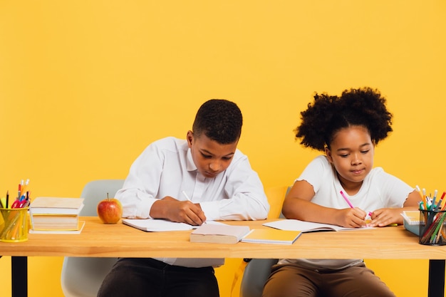 Happy african american schoolgirl and mixed race schoolboy sitting together at desk and studying on yellow background Back to school concept