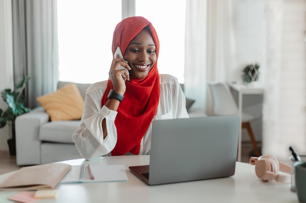 Happy african american woman in hijab working on laptop at home office and talking on smartphone with client