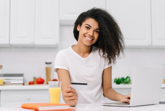 Happy African American woman shopping online, ordering food, standing on kitchen. Portrait of beautiful hipster girl using laptop, holding credit card, making payment, working from home